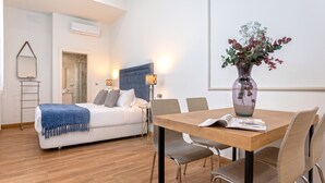 Een moderne kamer met een bed, een eettafel met stoelen, een vaas met bloemen en een klein bijzettafeltje met een lamp.
