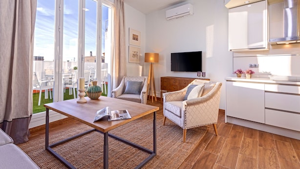 Un salon moderne avec une table basse en bois, deux fauteuils, un canapé et un coin cuisine doté de meubles blancs et d’un téléviseur.
