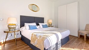 Un cuarto de hotel ordenado con una cama grande, dos mesitas de noche con lámparas, un espejo redondo, un armario y una silla.