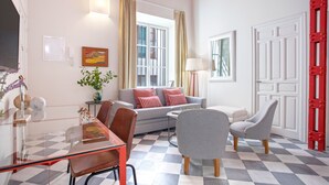 Un salon avec un sol à carreaux, un canapé, deux fauteuils, une table basse en verre et un téléviseur fixé au mur.