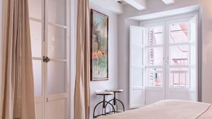 A bedroom with a bed, a small table, a chair, a painting, and a window with a view of a balcony. - Genteel Home Plaza de España II (Cadiz)