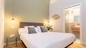 Ein ordentlich bezogenes Bett mit weißen Leinentüchern und grauen Kissen, ein Nachttisch mit zwei Vasen sowie eine hängende Lampenleuchte.