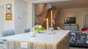 A modern kitchen with a central island, a dining area with chairs, a staircase, and a living room with a sofa and TV.
