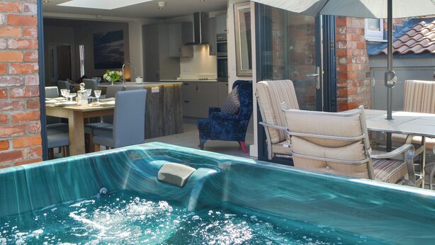 A swimming pool with a glass enclosure, a dining area with chairs and a table, and a kitchen visible through the glass doors.