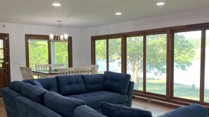 Ein modernes Wohnzimmer mit einem großen, blauen Ecksofa, einem Esstisch mit Stühlen und großen Fenstern mit Blick auf Grünflächen.