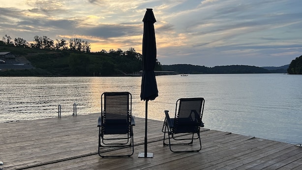Eine Holzterrasse mit zwei Liegestühlen und einem Sonnenschirm, mit Blick auf ruhiges Wasser, eine Küstenlinie und Bäume im Hintergrund.