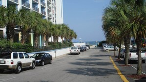 A street lined with parked cars, palm trees, and a tall building with balconies.
