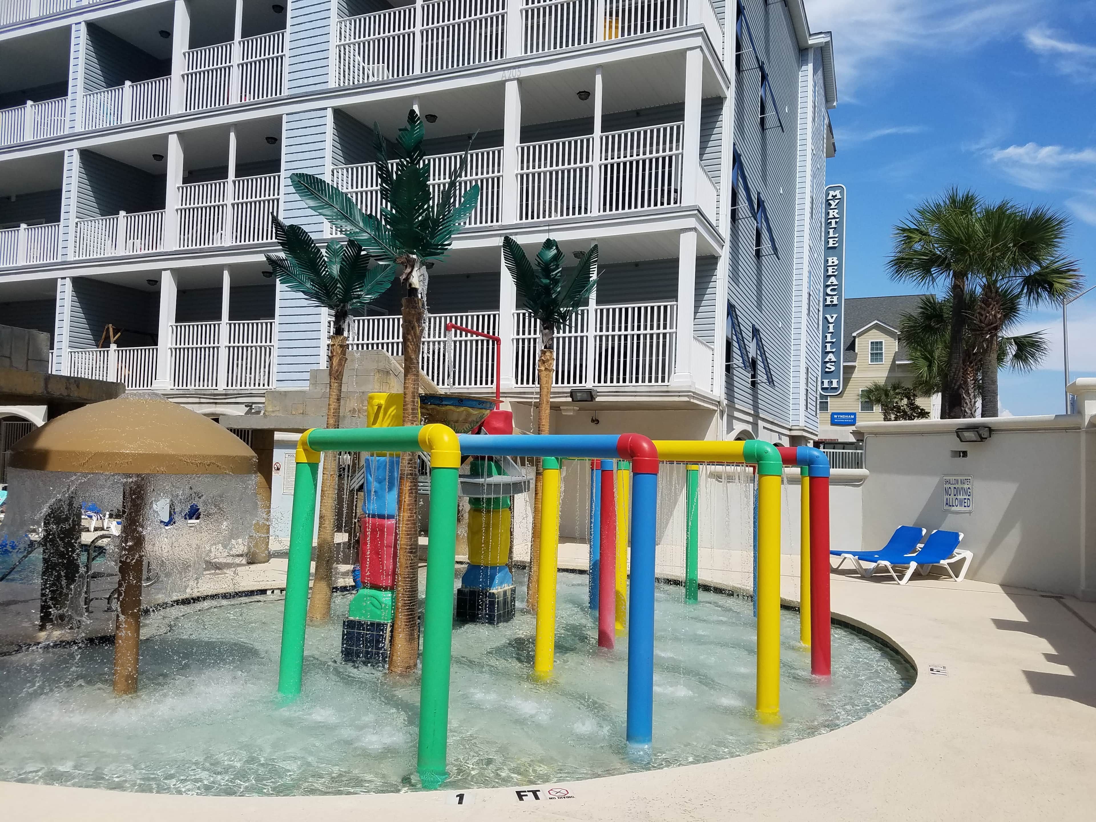A water play area with colorful structures, a pool, and a building with balconies in the background.