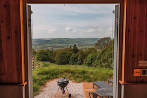 A wooden-framed door opening to a patio with a grill, outdoor furniture, and a scenic view of hills and trees. - Peak District Shepherds Hut with incredible views (Derbyshire)