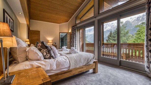 A bedroom with a wooden bed, a nightstand, a lamp, and a view of mountains through large windows. - Chalet Birdie + Mazot: elegant and spacious property in Les Houches (Les Houches)