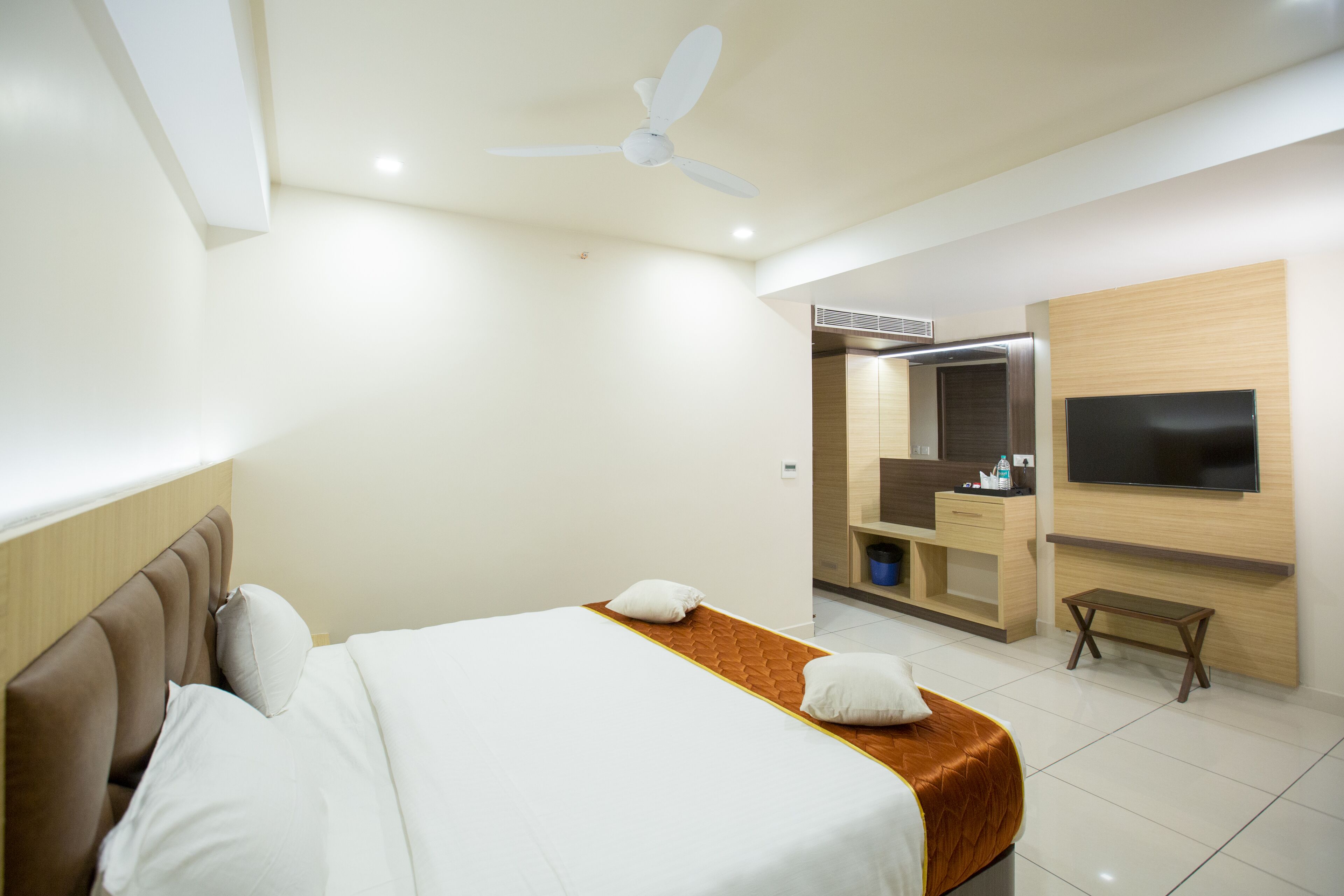 A hotel room with a bed, a ceiling fan, a TV mounted on the wall, and a small wooden bench.