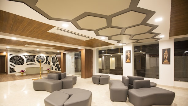 A modern lobby with a geometric ceiling design, a seating area with gray sofas, and a reception desk with a decorative wall. - Hotel Ark (Kumbakonam)