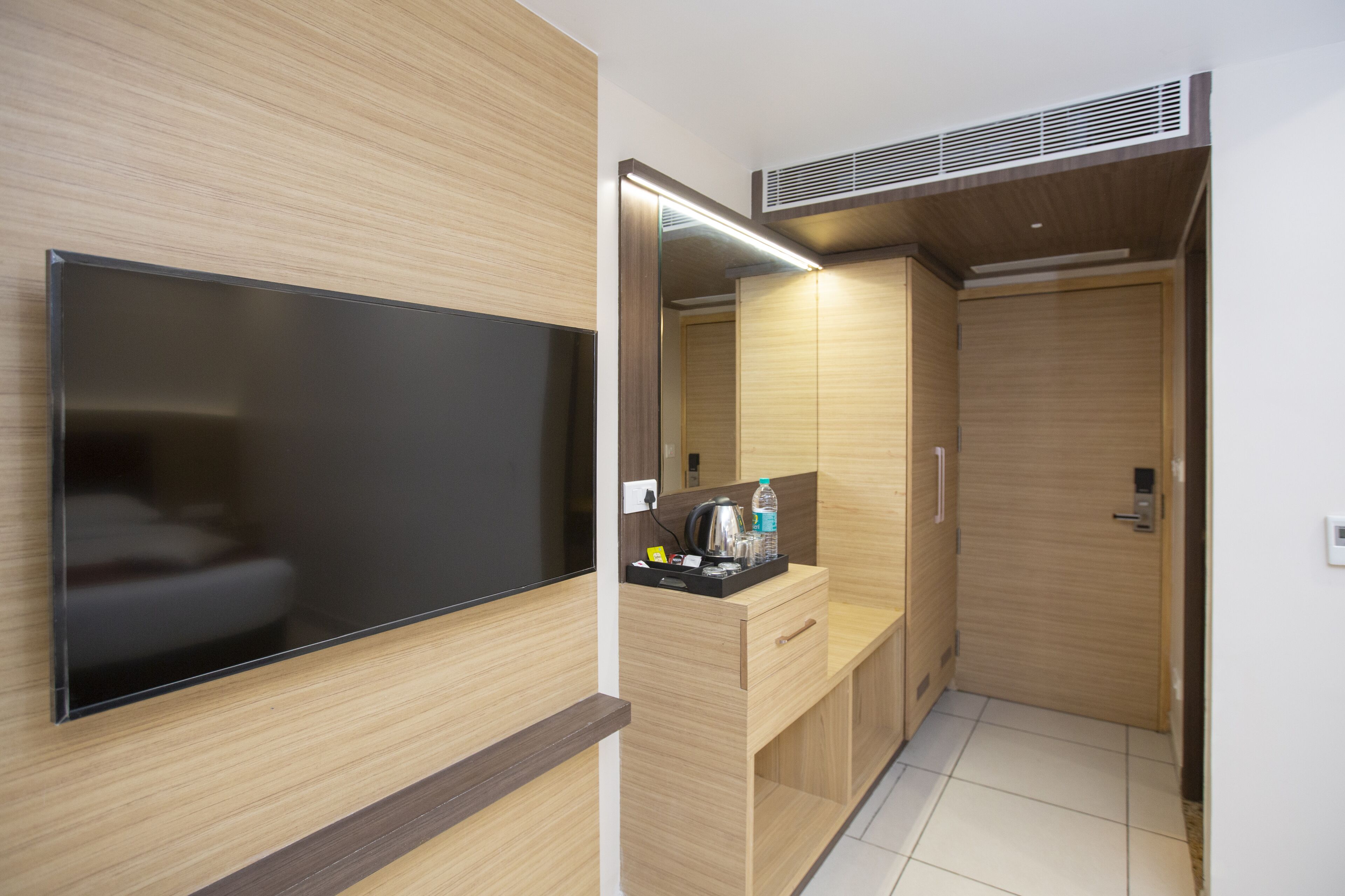A modern hotel room with a flat-screen TV, a wooden vanity with a mirror, a sink, and a small countertop with toiletries.