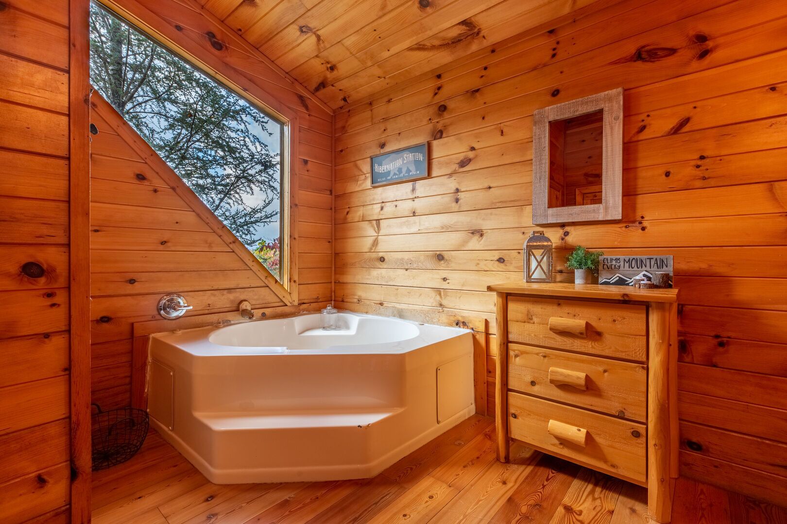 A wooden cabin bathroom with a large bathtub, wooden vanity, and a window offering a view of trees.