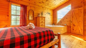 A cozy cabin room with a large bed, a wooden ceiling, a bathroom with a bathtub, and a large window.
