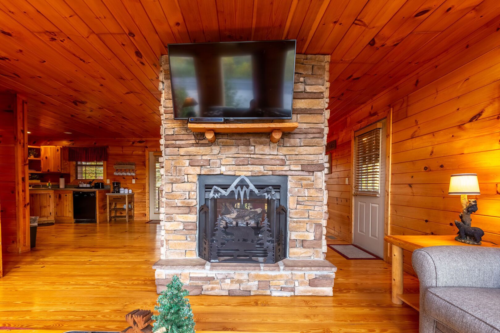A cozy cabin interior with a stone fireplace, a flat-screen TV mounted above it, wooden floors, and a dining area visible in the background.