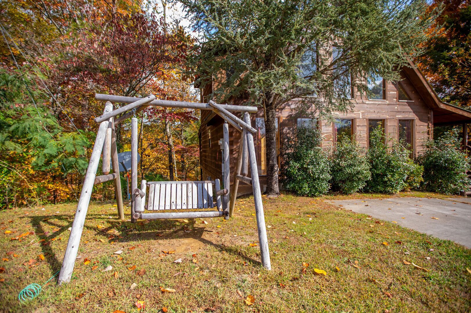 Een houten schommel in een park met herfstbladeren, een stenen gebouw op de achtergrond en een heldere lucht.