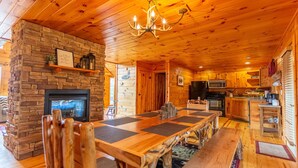 A cozy cabin interior with a stone fireplace, wooden dining table, and a kitchen area.
