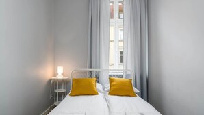 Una habitación pequeña con una cama individual, estructura de metal blanco, dos almohadas amarillas, ropa de cama blanca, una mesita de noche con lámpara y una ventana con cortinas.