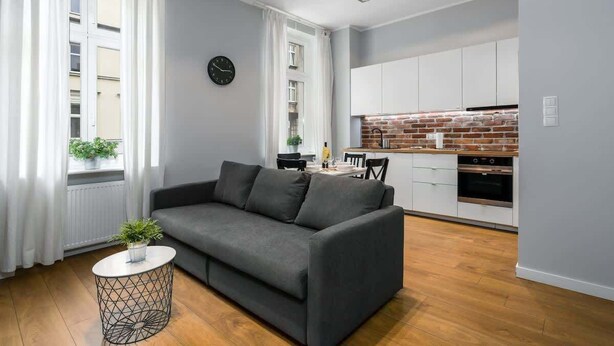 Un apartamento moderno con un sofá gris, una mesita redonda con una planta, una cocina con armarios blancos y un salpicadero de ladrillo, y una ventana con cortinas blancas.