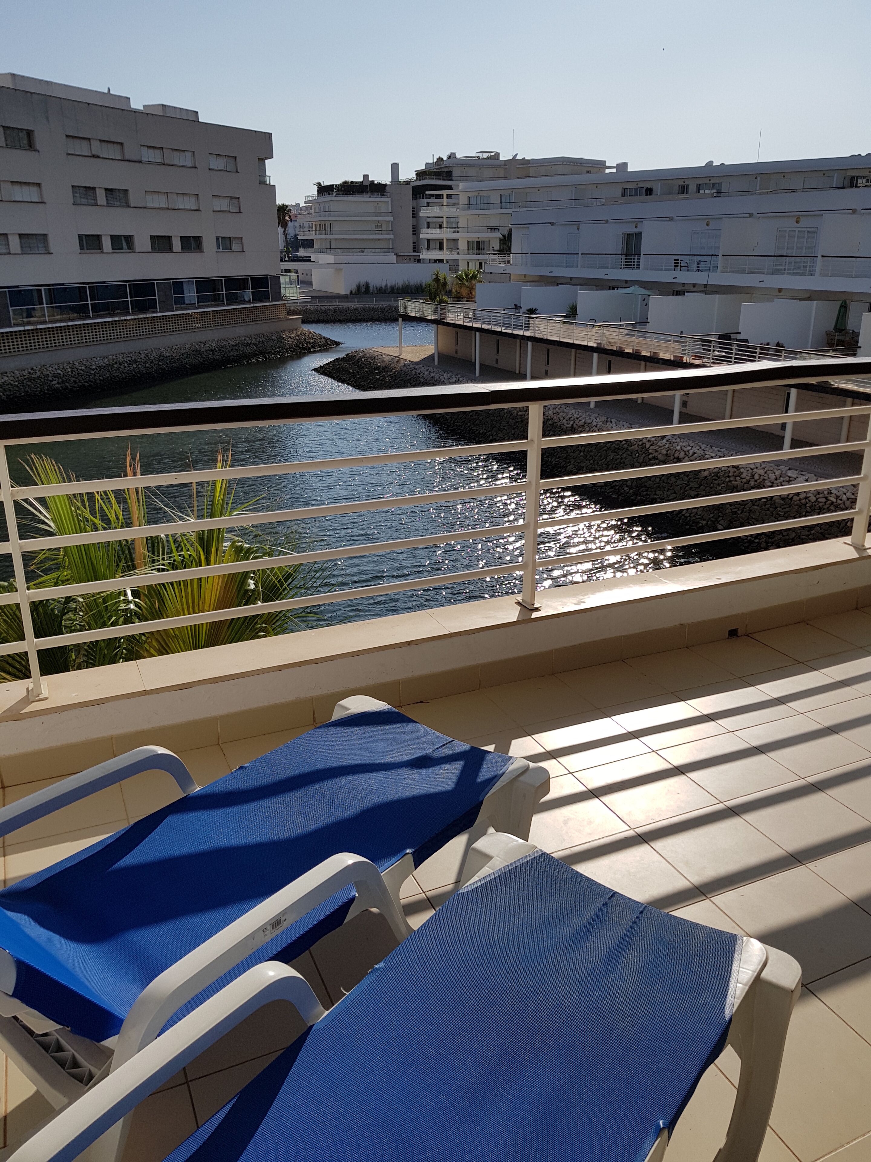 Ein Balkon mit zwei blauen Liegestühlen, einer kleinen Wasserspende und Blick auf moderne Gebäude.