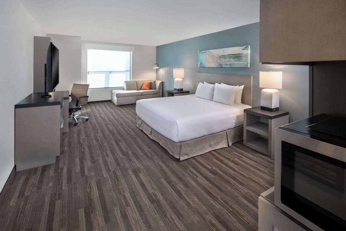 A modern hotel room with a large bed, a desk with a flat-screen TV, a sofa, and a dining table.