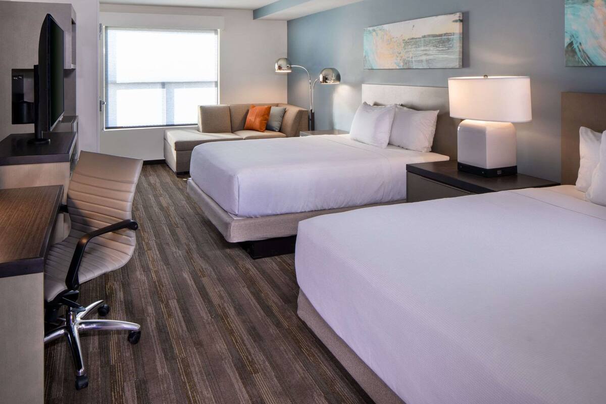 A hotel room with two beds, a desk, a chair, a TV, and a window.