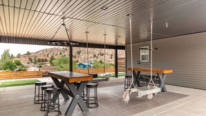 Un patio al aire libre con una mesa de madera y taburetes de bar, una hamaca colgante y vistas a un paisaje montañoso.