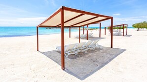 A beach with white lounge chairs under a shaded area, a clear blue sky, and the ocean in the background. - Breathless 4You, Oceanfront Penthouse (Cap Cana)