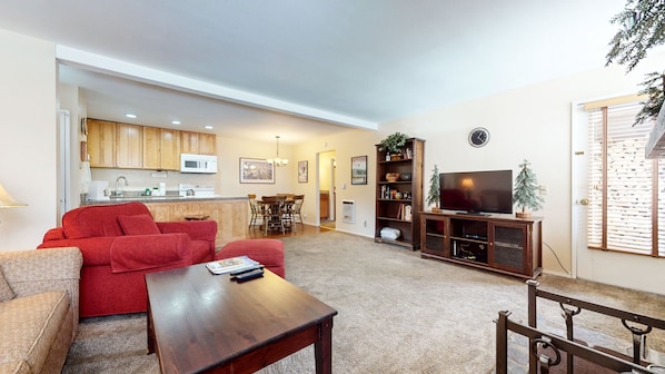 A living room with a red sofa, a wooden coffee table, a TV on a wooden stand, and a kitchen in the background.