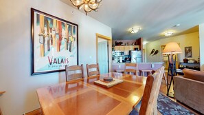 A dining area with a wooden table and chairs, a framed artwork on the wall, and a kitchen in the background.