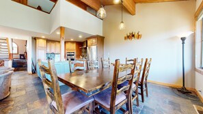Un vaste espace de salle à manger avec une grande table et des chaises en bois, une cuisine à l’arrière-plan et un escalier menant à un niveau supérieur.