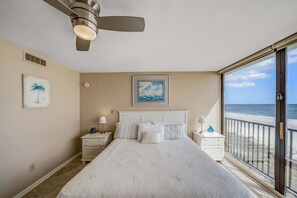 Une chambre avec un grand lit, un ventilateur de plafond, une table de chevet avec une lampe, un tableau représentant un palmier et une vue sur la plage, visible à travers une porte coulissante.