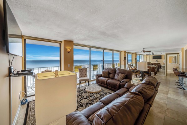 A spacious living area with a large window overlooking the ocean, a brown leather sofa, and a dining area with chairs and tables.