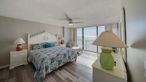 A bedroom with a bed, nightstands, a green lamp, a ceiling fan, and a view of the ocean through large windows.