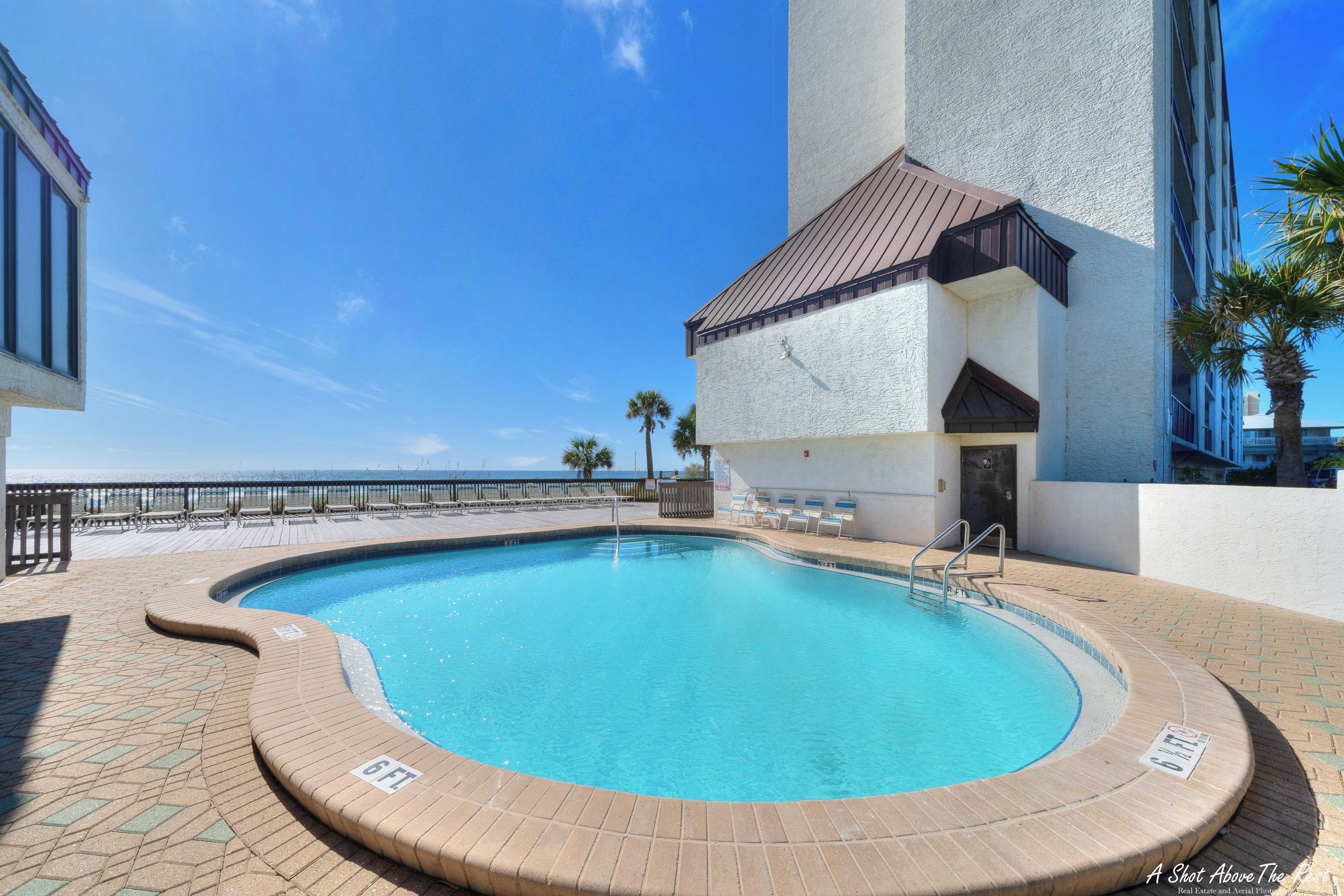 Une piscine dont la profondeur est indiquée à 6 pieds, entourée d’une terrasse en briques et d’une rambarde, et située à côté d’un bâtiment à la façade blanche et au toit brun.