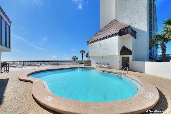 A swimming pool with a 6ft depth marker, surrounded by a brick deck and a railing, adjacent to a building with a white facade and a brown roof.