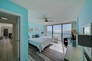 A bedroom with a large bed, a ceiling fan, a balcony with a view of the ocean, and a dining area with a table and chairs.