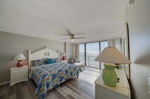 A bedroom with a bed, nightstands, a green lamp, a ceiling fan, and a view of the ocean through large windows.
