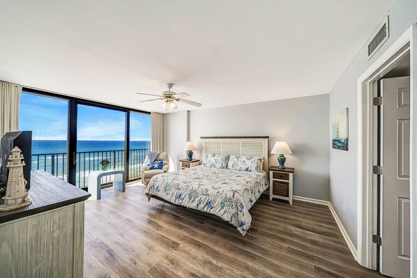 A bedroom with a large bed, a ceiling fan, a nightstand with a lamp, a bench by the window, and a view of the ocean. - Seachase E801 Enjoy Incredible Views, FREE Beach Service, FREE Golf & Perks! (Panama City Beach)