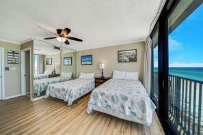 A hotel room with two beds, a ceiling fan, a large window with a view of the beach, and a wooden floor.