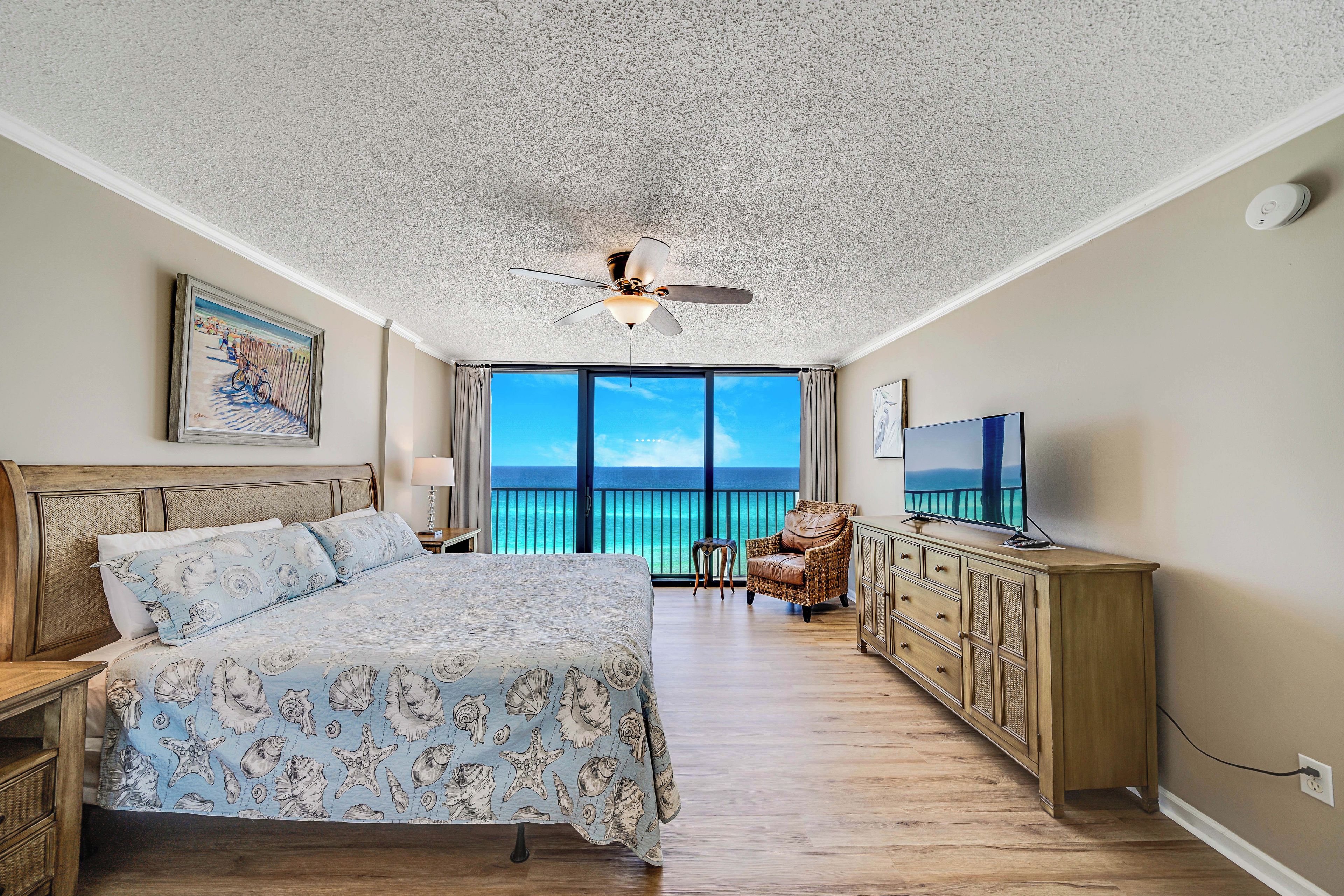 A bedroom with a large bed, a ceiling fan, a TV, a dresser, and a view of the ocean.