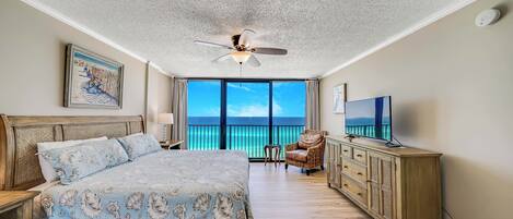 A bedroom with a large bed, a ceiling fan, a TV, a dresser, and a view of the ocean.