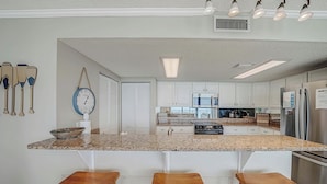 A kitchen with a central island, white cabinets, stainless steel appliances, and wooden stools. - FREE Beach Service for 2, FREE Daily Golf & Perks! Beachfront Seachase E802 (Panama City Beach)