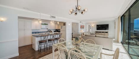 Una cocina moderna con armarios blancos, una isla central y un comedor con mesa de vidrio y sillas de mimbre.