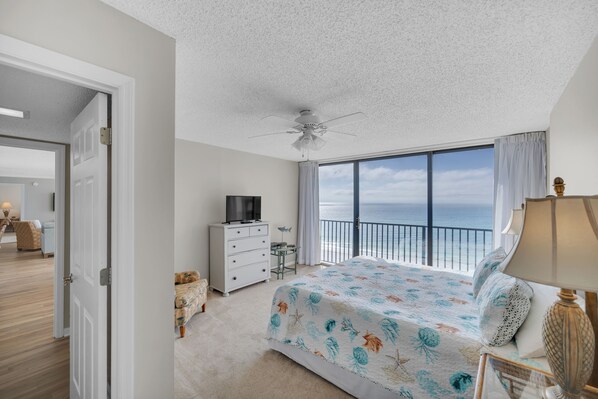 A bedroom with a bed, a ceiling fan, a dresser, a TV, and a view of the ocean.