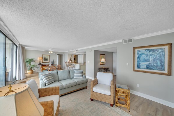 A spacious living room with a light-colored sofa, a wicker chair, and a dining area in the background.