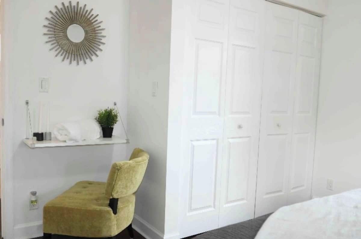 Een kamer met een witte kast, een kleine stoel, een spiegel aan de muur en een plank met toiletartikelen.