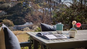 A table with a book, a mug, and a vase with flowers, overlooking a coastal area with houses and trees. - 4 BR (Sleeps 7) in Salcombe overlooking beach (Salcombe)