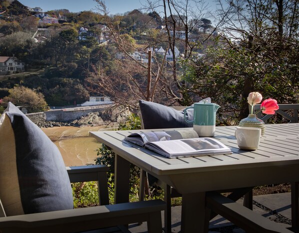 A table with a book, a mug, and a vase with flowers, overlooking a coastal area with houses and trees.